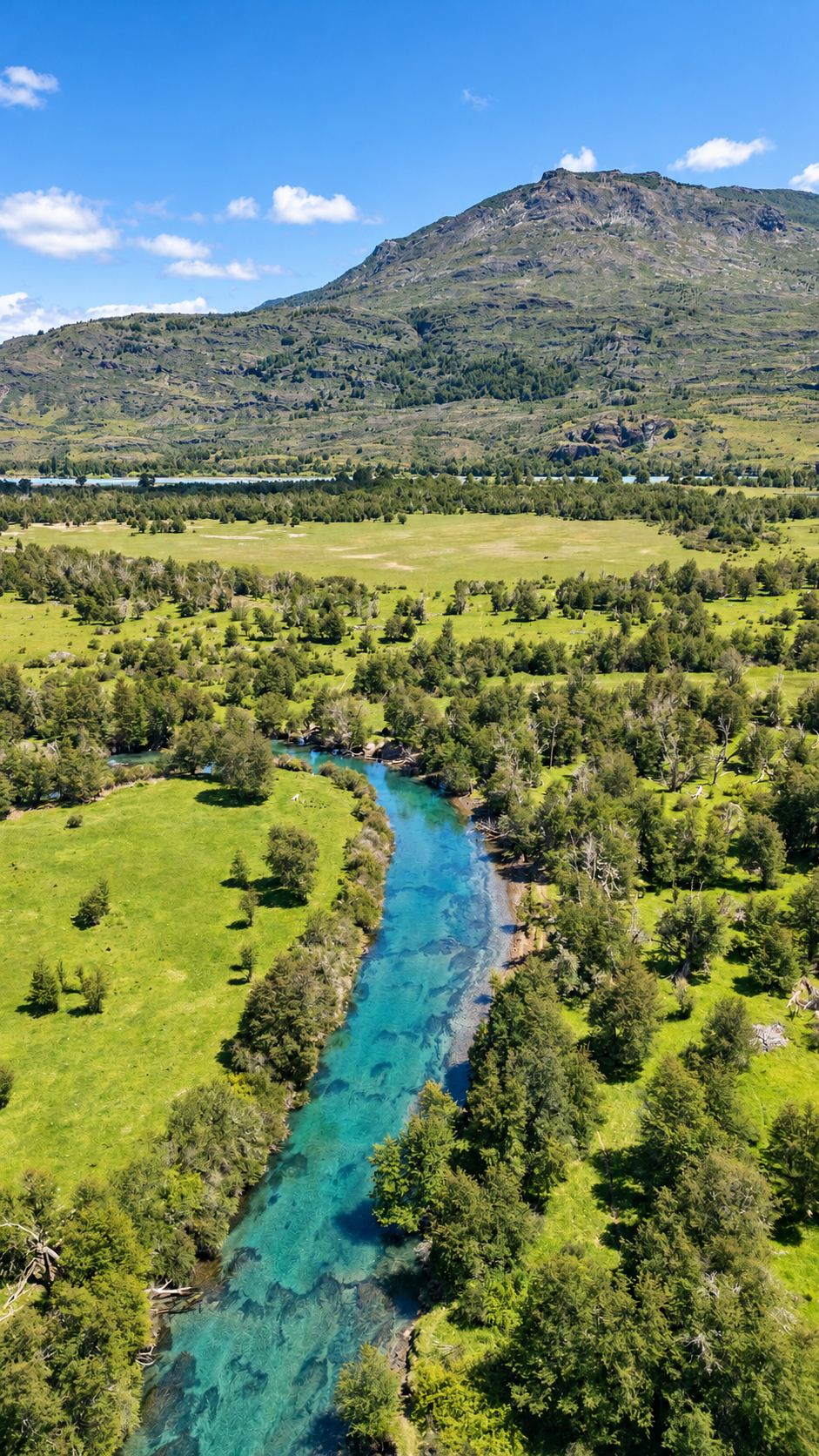 Aerial view of a turquoise river winding through meadows toward a mountain